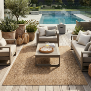 A large natural sisal outdoor area rug in a neutral tan colour, styled in a modern patio setting with a wooden coffee table, beige and grey outdoor lounge chairs, and a swimming pool in the background.